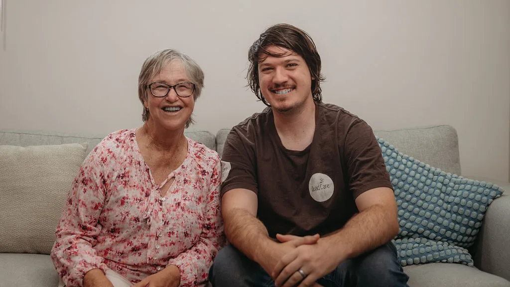 An older woman and a young man sitting together on a lounge at home, both smiling at the camera